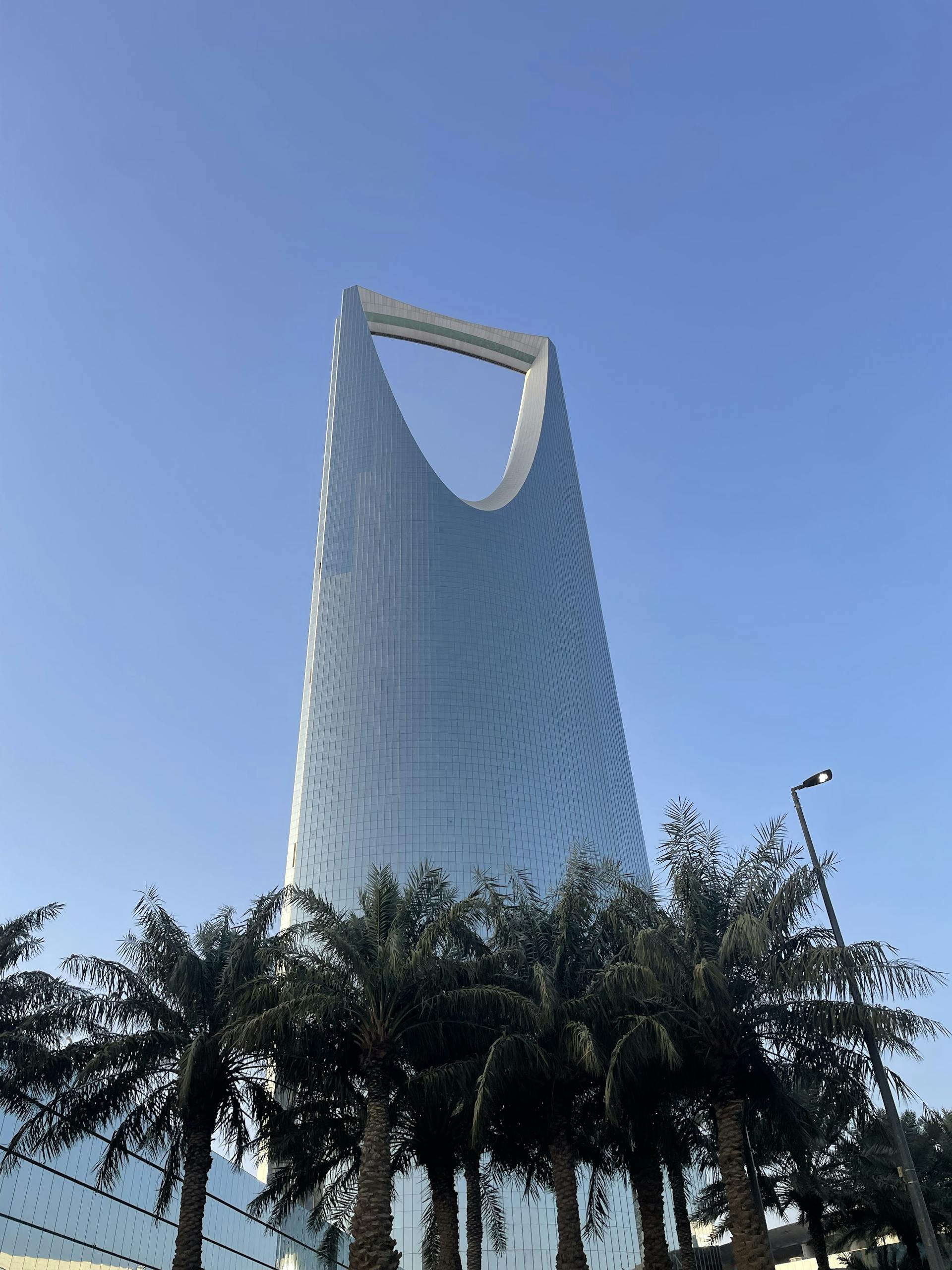 Tall skyscraper with unique design in Riyadh, framed by palm trees against a clear sky.